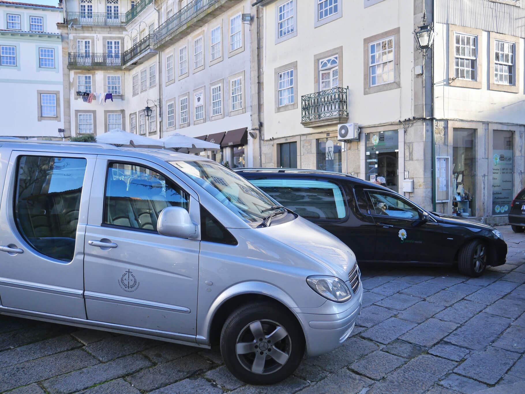 Uma van prateada e um carro funerário preto estacionados em uma rua de paralelepípedos em frente aos prédios.