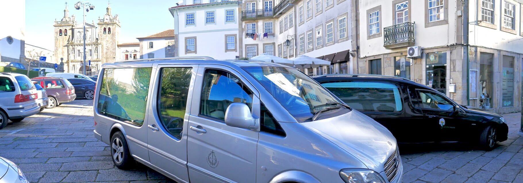 Uma van funerária prateada estacionada em uma rua de paralelepípedos, com um carro funerário preto por perto.