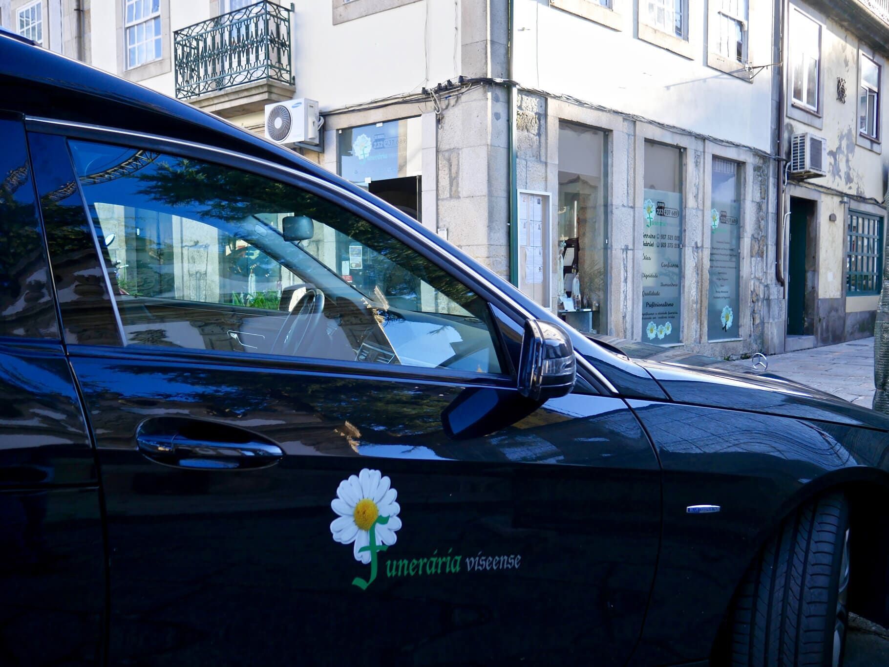Carro funerário preto com logotipo de margarida, estacionado em frente a um prédio com lojas.