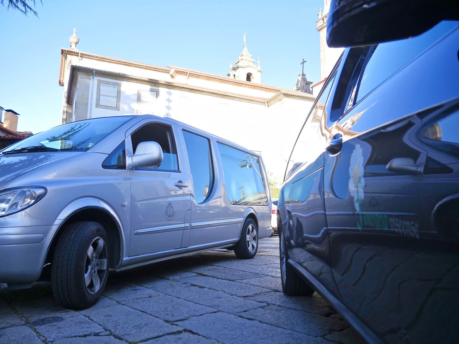 Van prateada e carro preto estacionados em rua de paralelepípedos, prédio ao fundo.