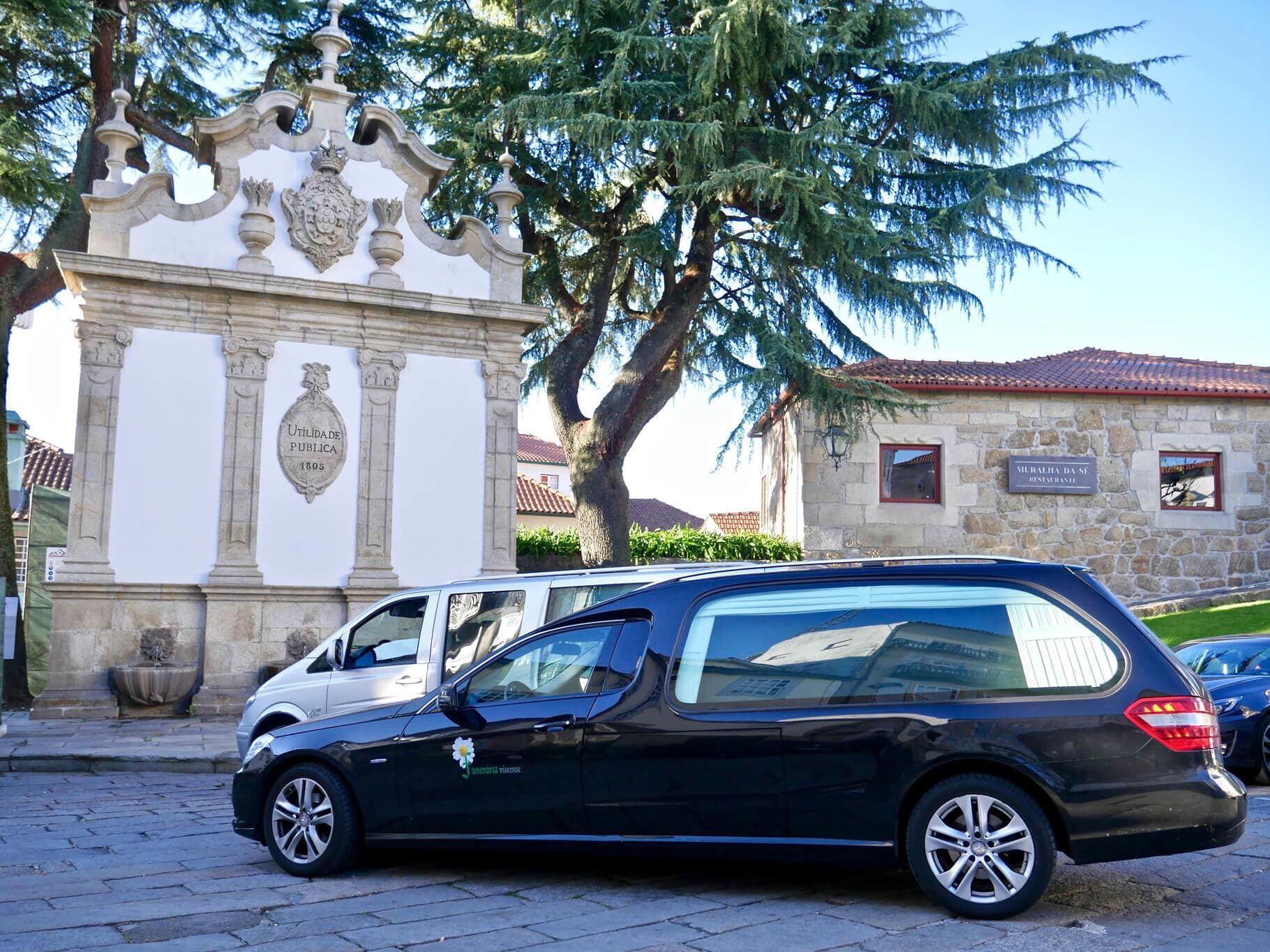 Carro funerário preto estacionado perto de uma fonte de pedra ornamentada em uma praça.