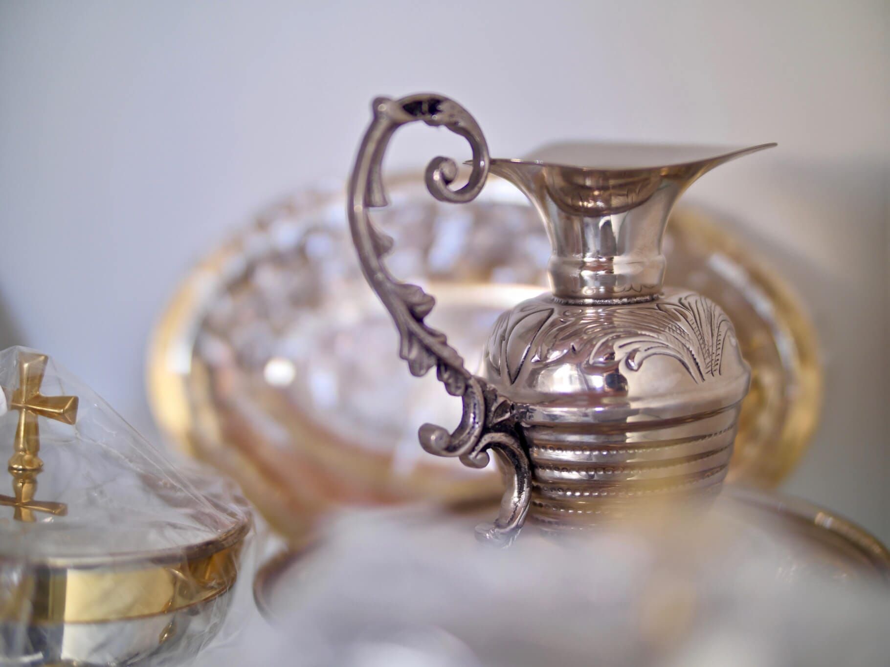 Jarro de prata com alça ornamentada, desenhos gravados e um prato redondo atrás.