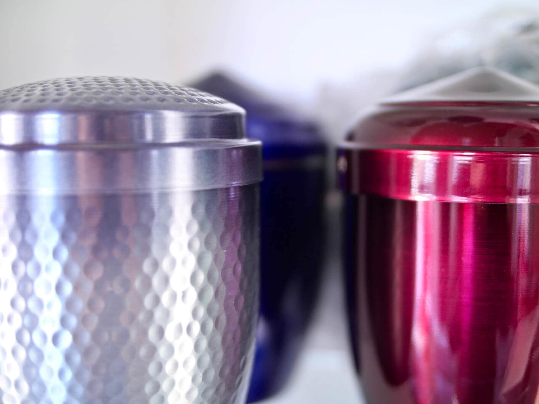 Três urnas coloridas: prata, azul e vermelha, em foco suave, provavelmente para cinzas.