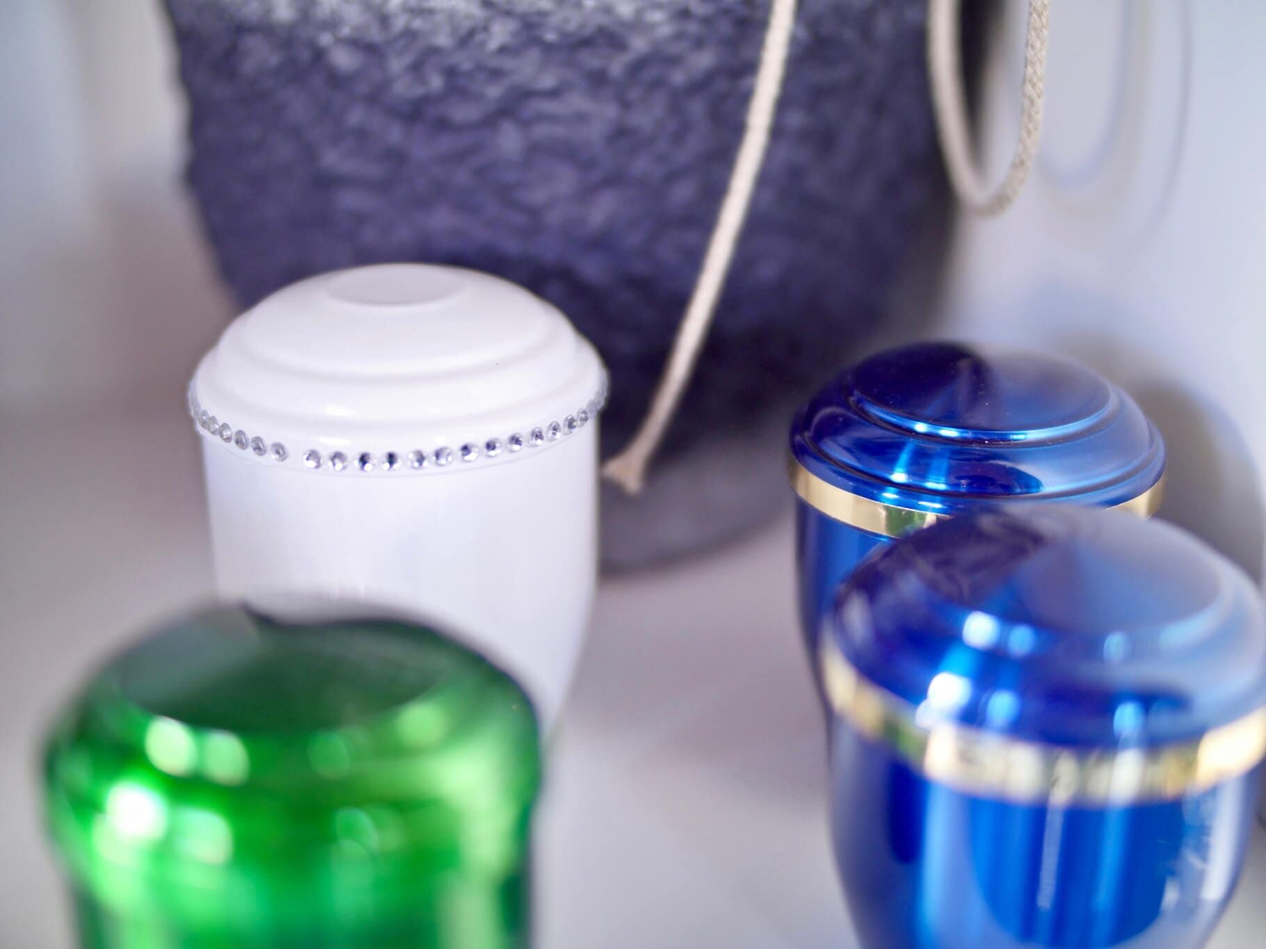 Quatro urnas coloridas em uma prateleira: branca, azul e verde. Ao fundo, um vaso azul desfocado.