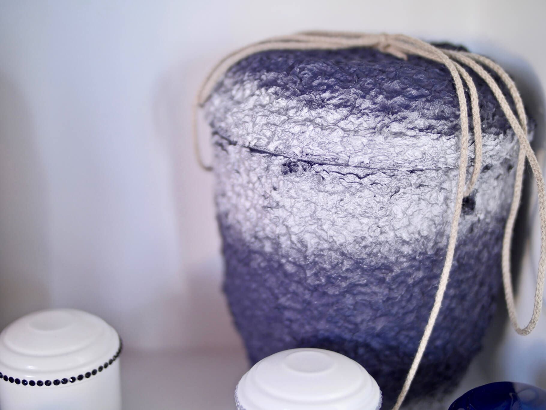 Urna de feltro azul e branca com corda, em uma prateleira com potes decorativos brancos.