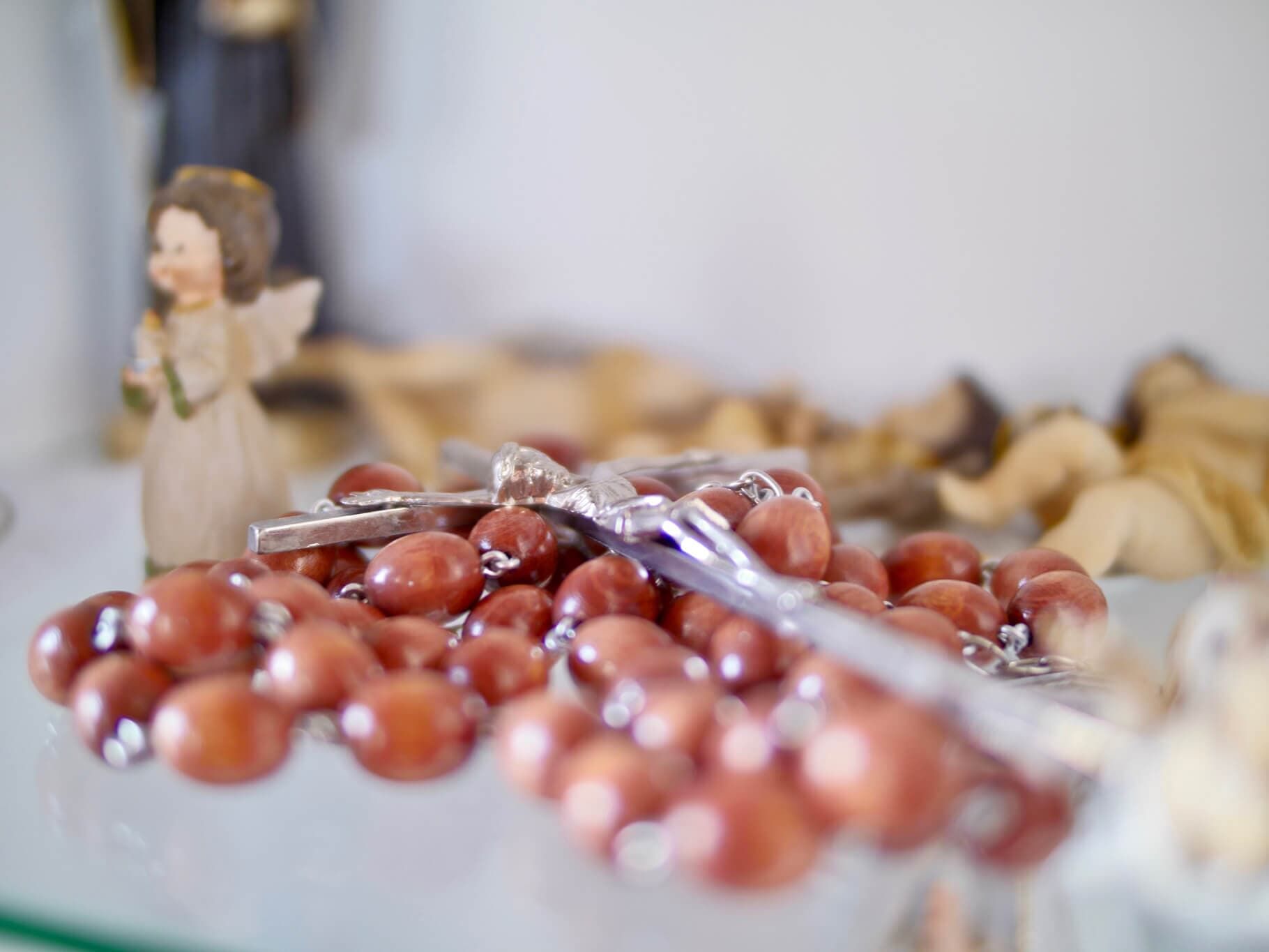 Rosário com contas marrons e cruz prateada, com uma pequena estatueta de anjo ao fundo.