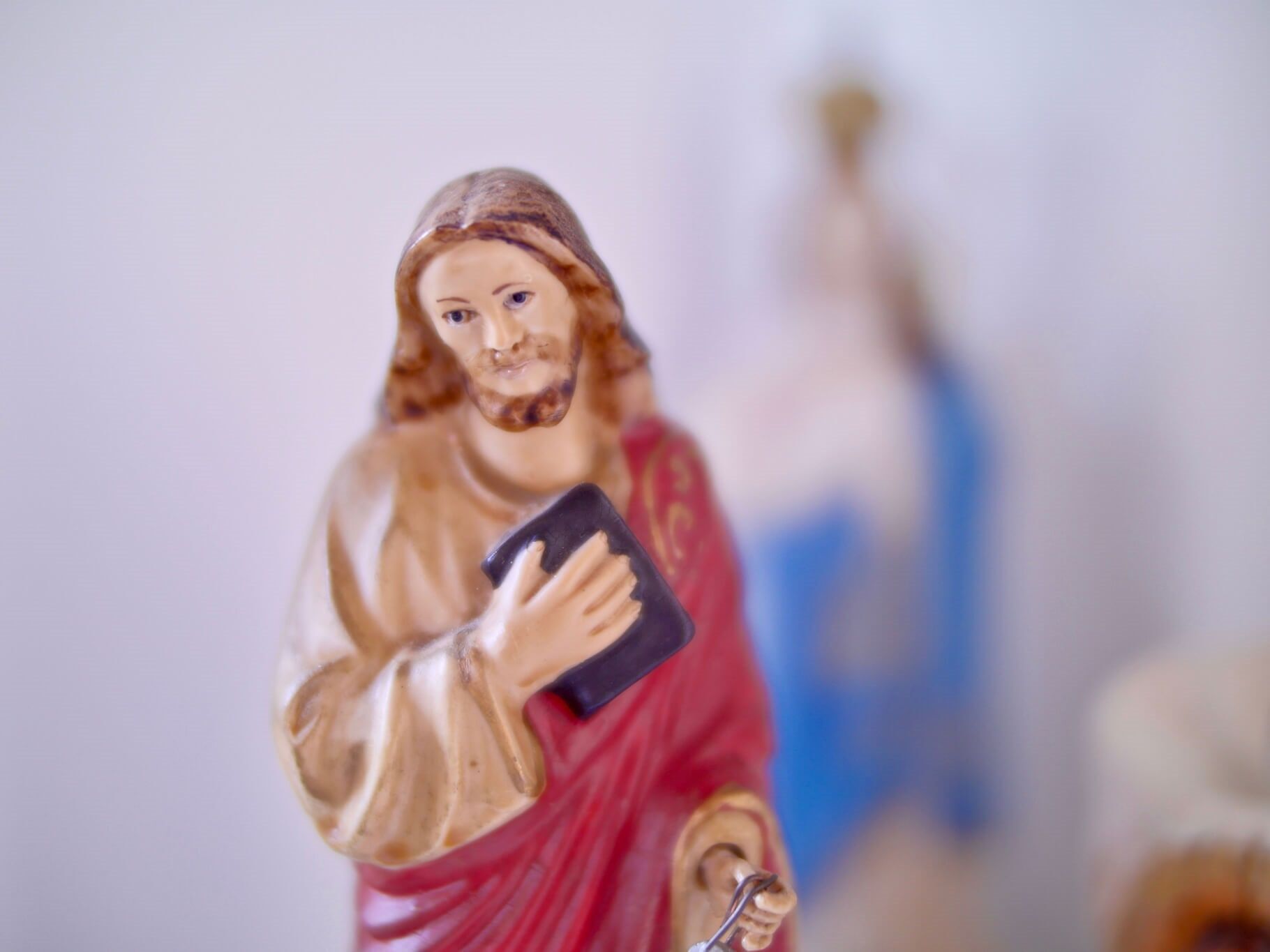 Estátua de Jesus segurando um livro, túnica vermelha, cabelo castanho, com uma estátua da Virgem Maria desfocada ao fundo.