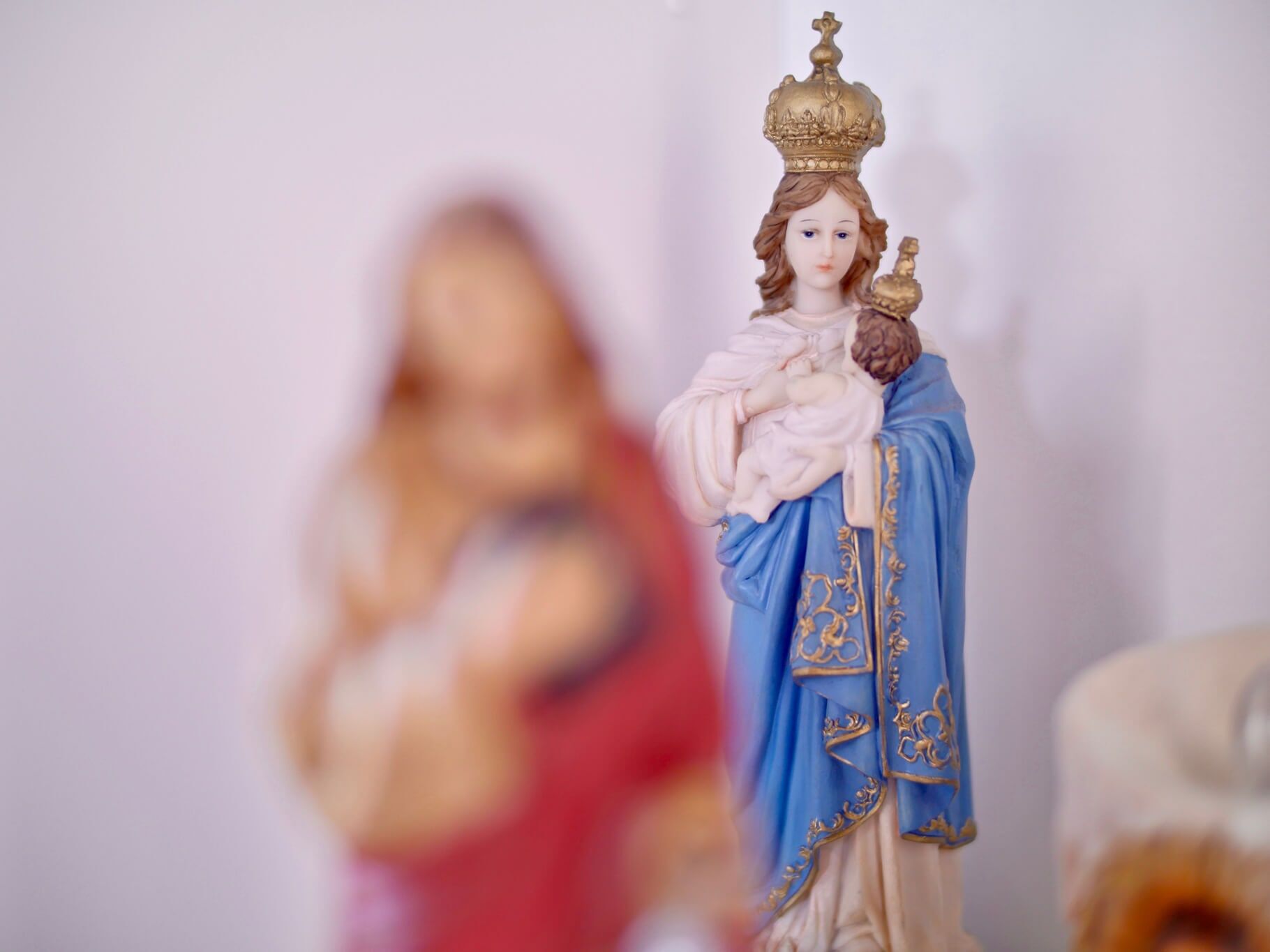 Estátua da Virgem Maria em túnica azul, segurando o menino Jesus, coroa de ouro, fundo branco.