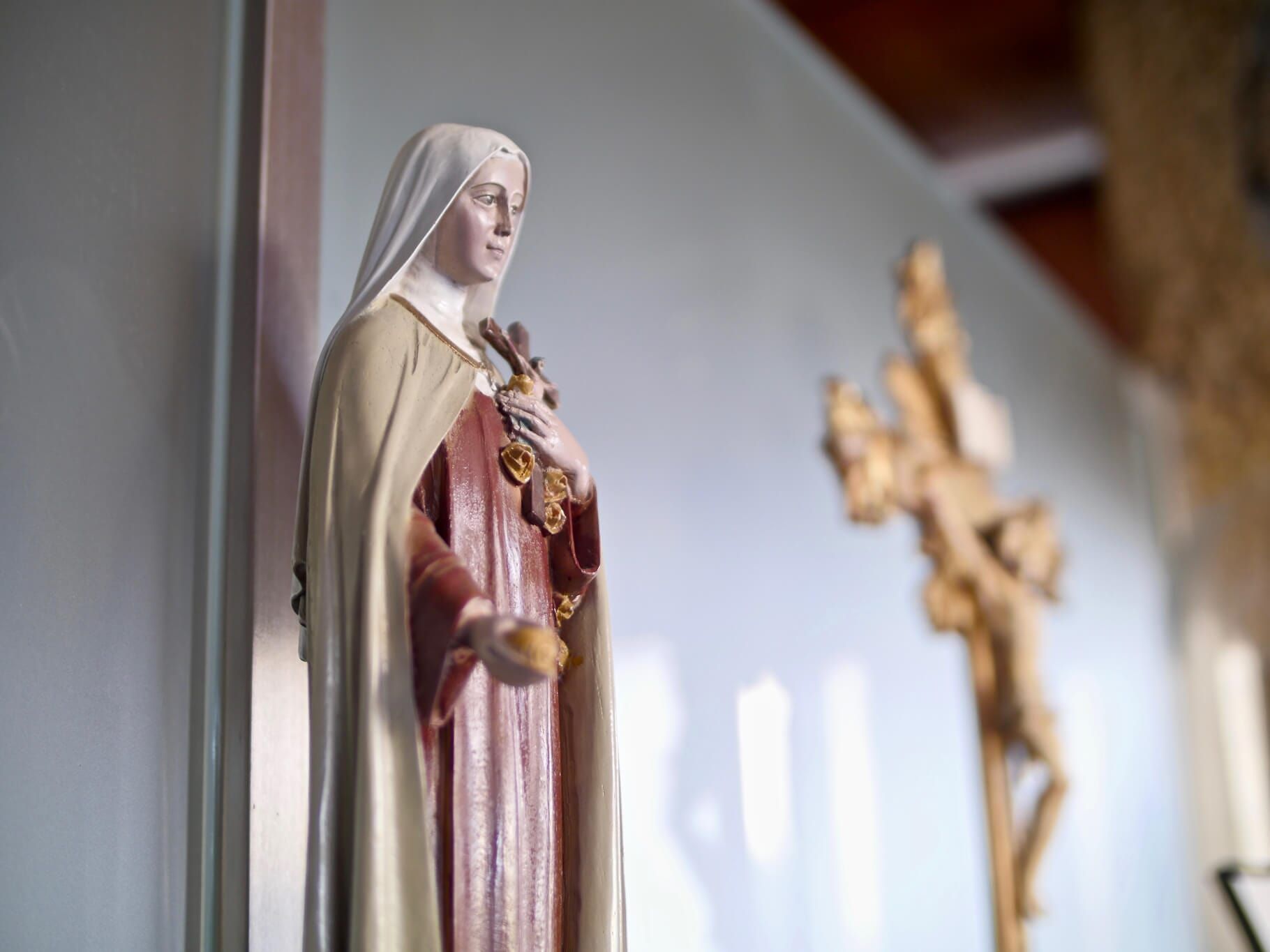 Estátua de um santo segurando um coração, com um crucifixo ao fundo. Tons de branco, marrom e vermelho.