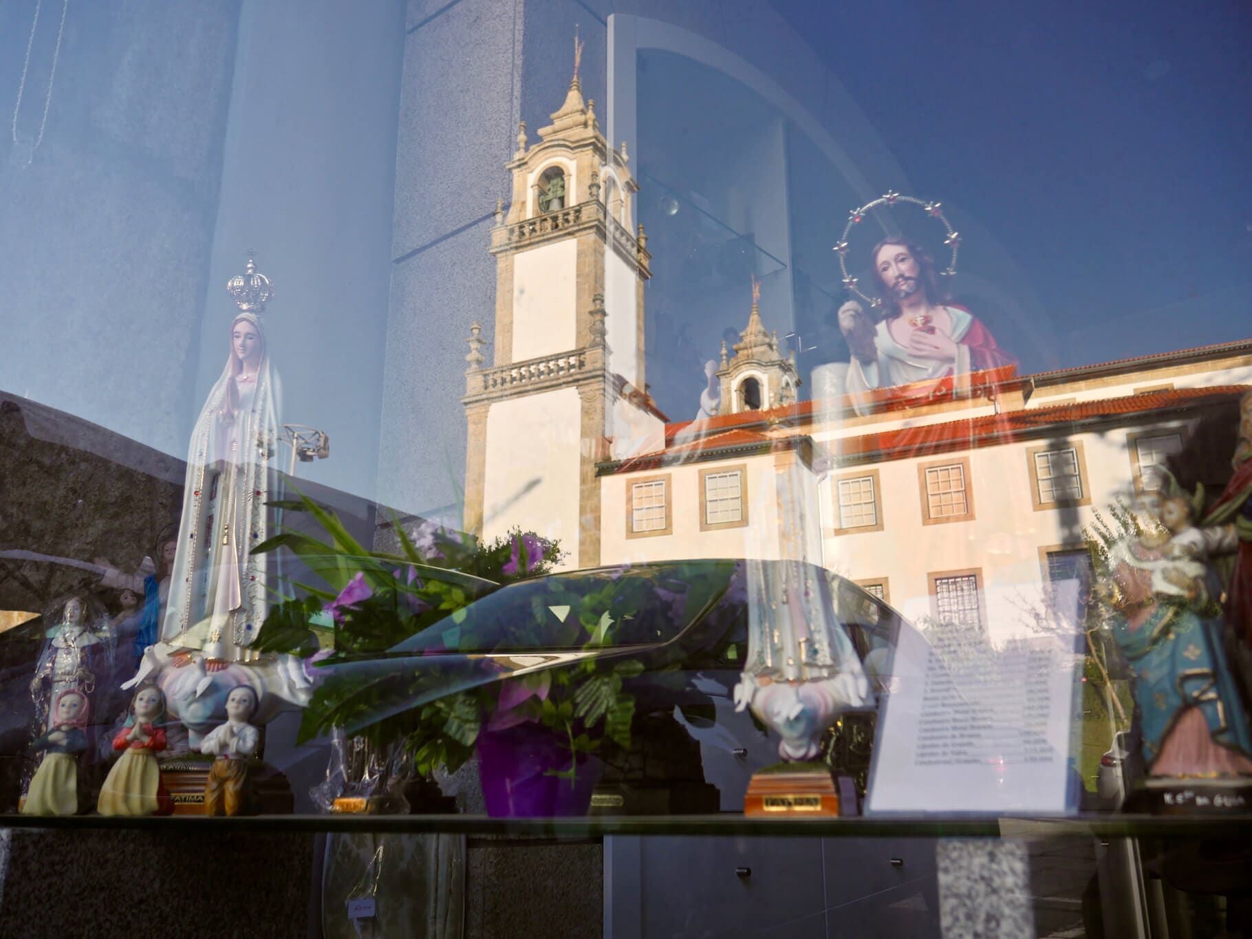Exposição de estátuas religiosas, flores e reflexos de uma igreja com uma torre.