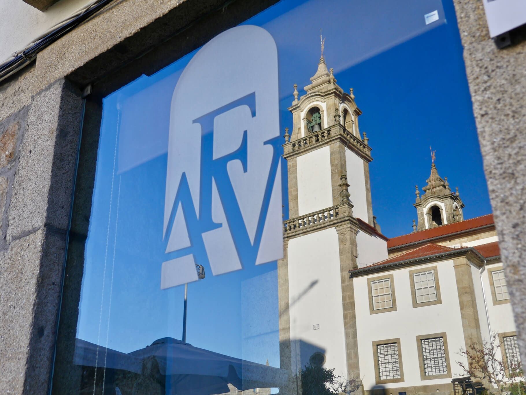 Janela com grande logotipo branco da AFV refletindo uma igreja branca com torres contra um céu azul.
