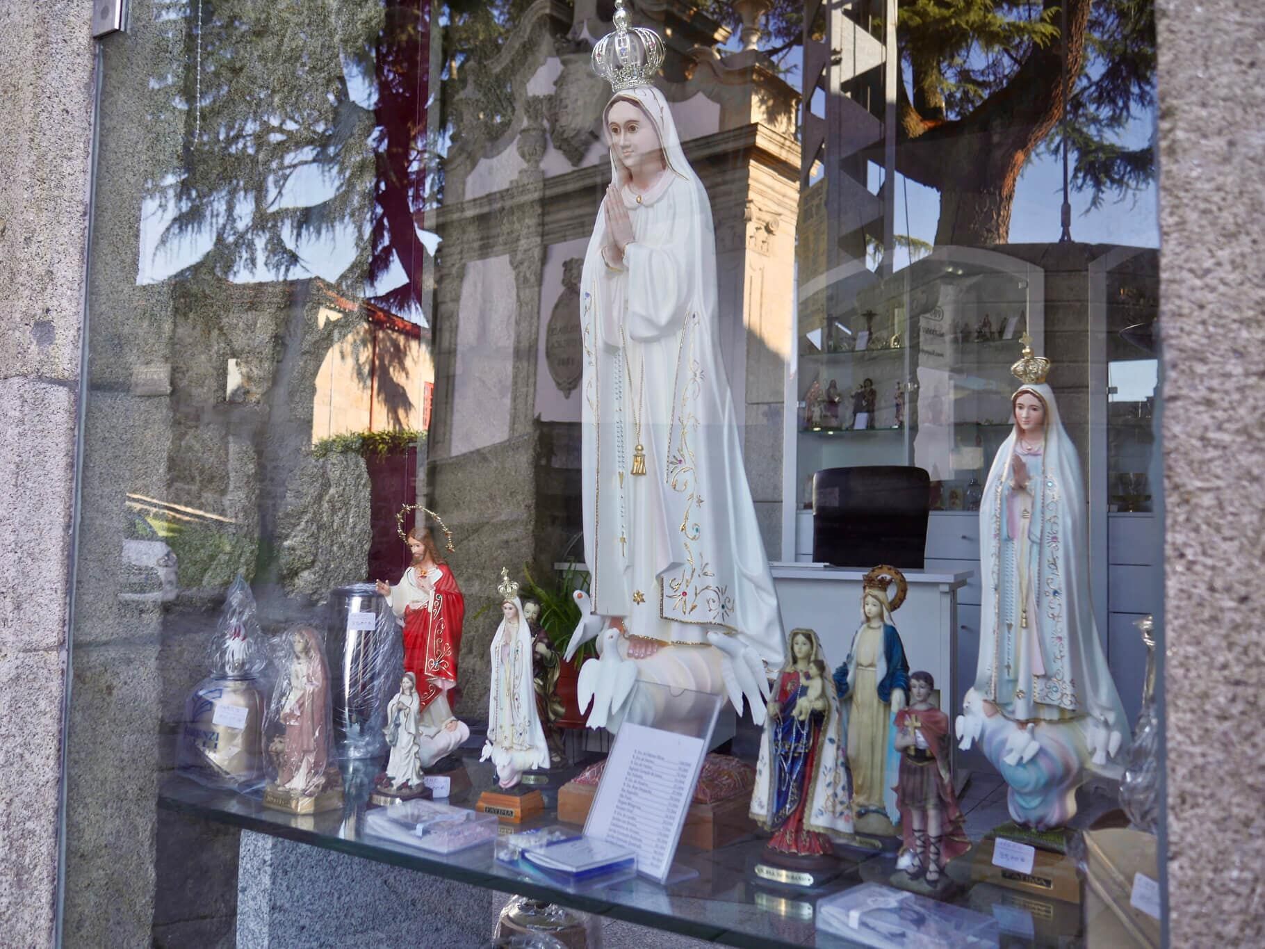 Estátuas da Virgem Maria expostas na vitrine de uma loja, com diversas cores e desenhos.