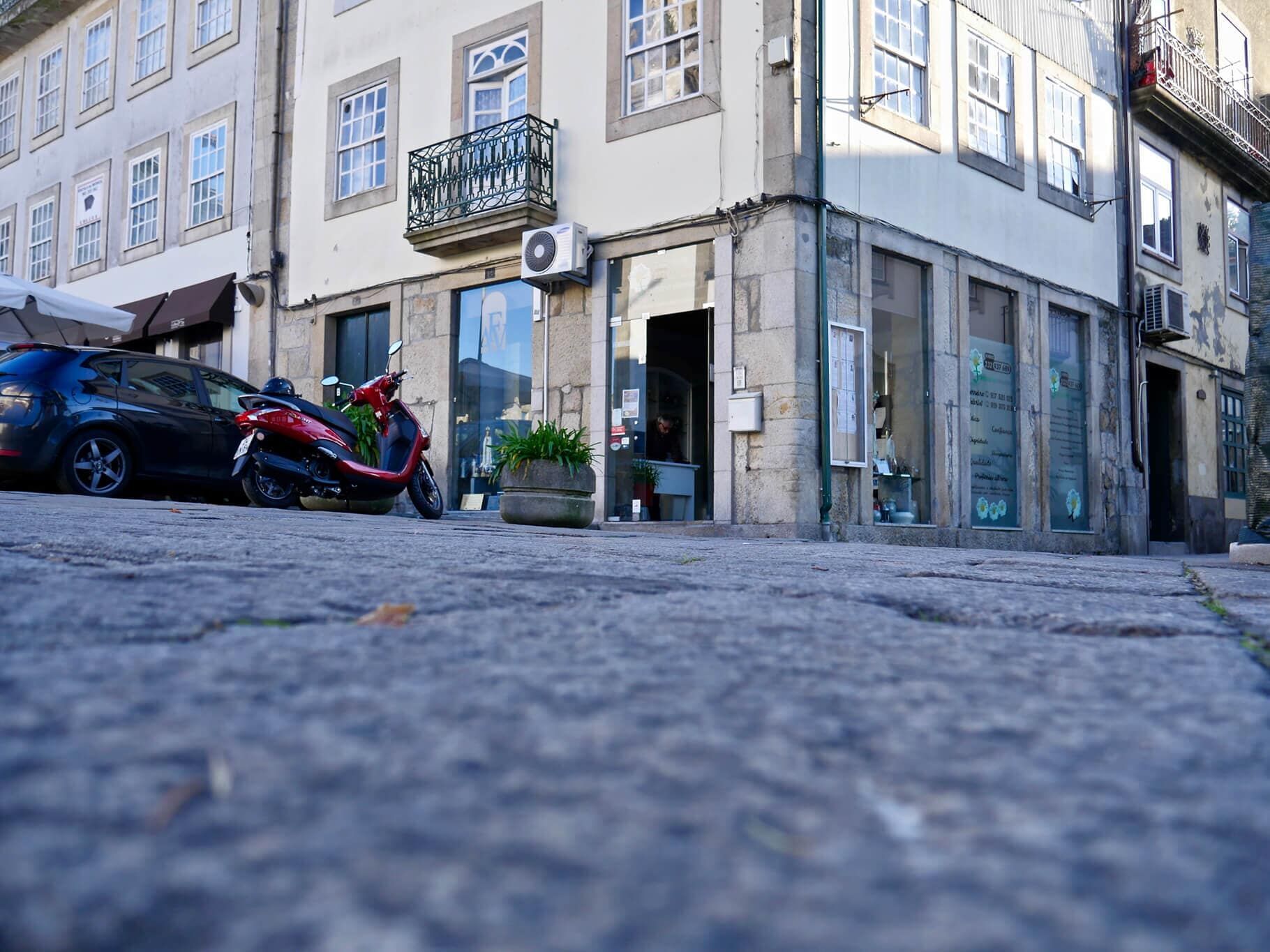Esquina com prédio, scooter e carro estacionado em rua de paralelepípedos.