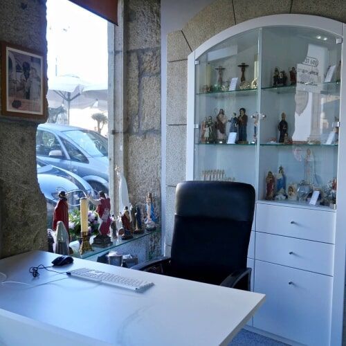 Escritório com escrivaninha, imagens religiosas e uma vitrine. Um carro é visível pela janela.