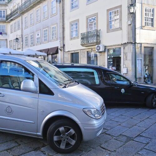 Uma van prateada e um carro funerário preto estacionados em uma rua de paralelepípedos em frente a prédios.