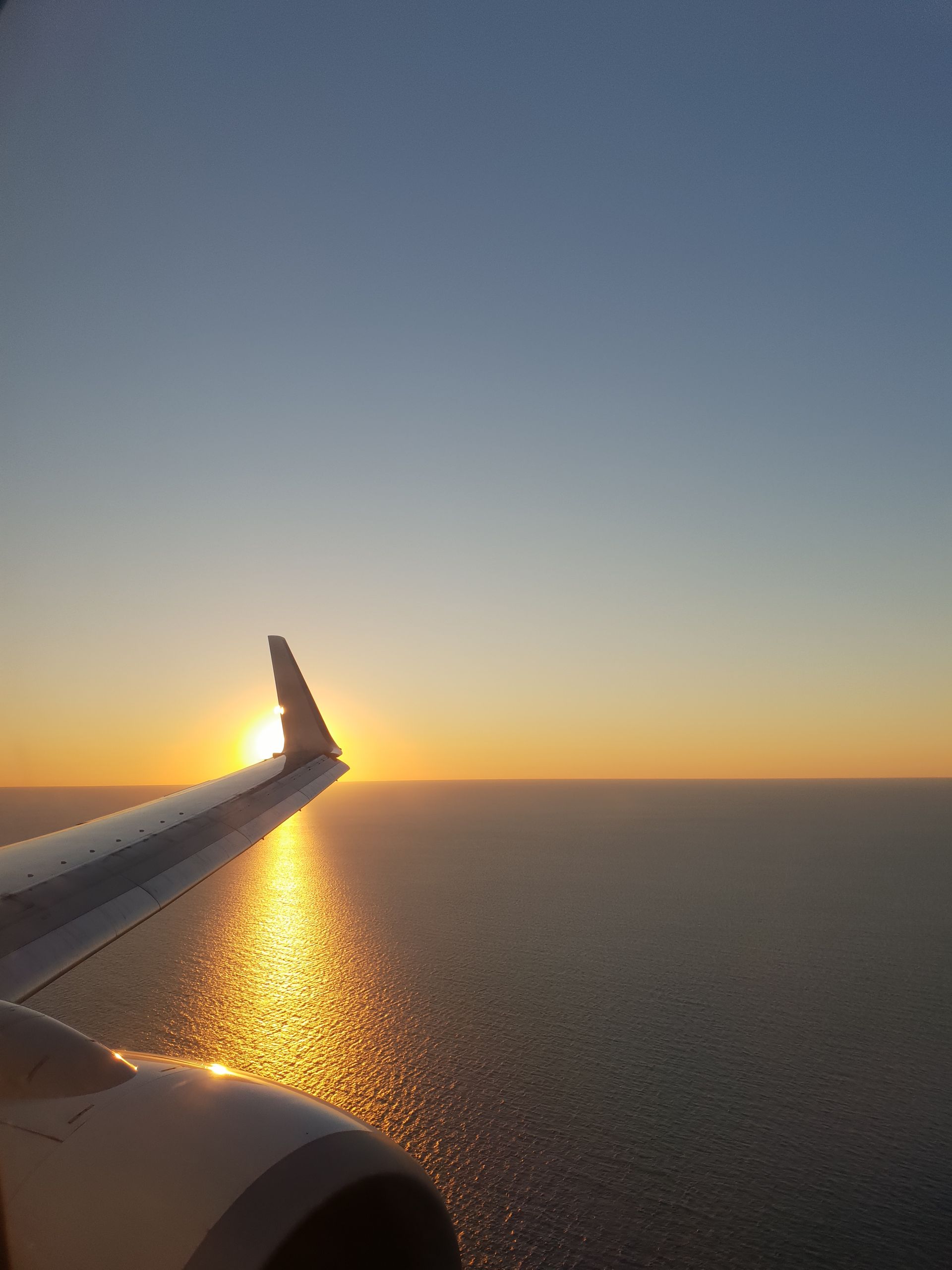 Aile d'avion au coucher du soleil au-dessus d'une mer calme, la lumière dorée se reflétant sur l'eau.