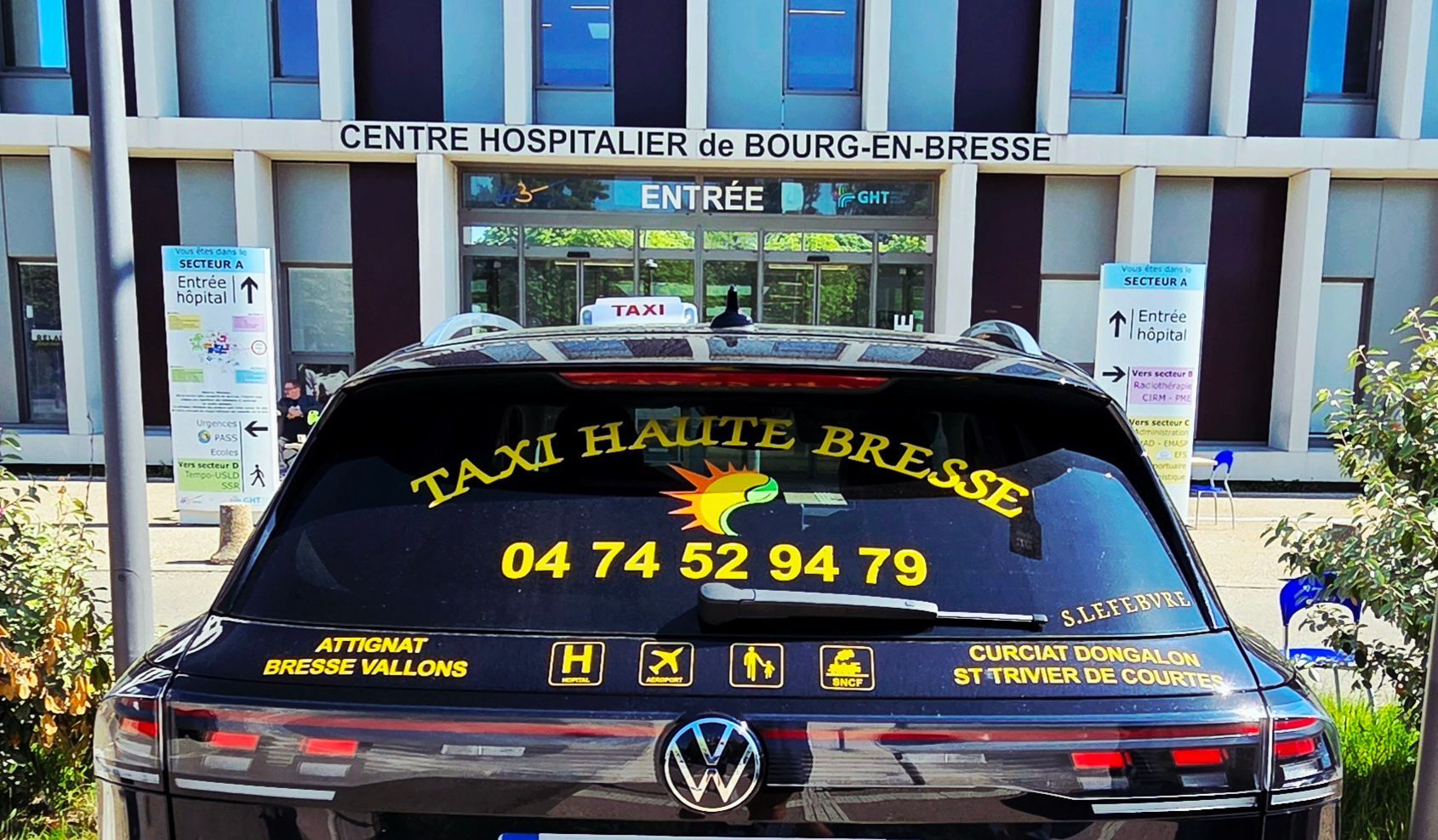 Un taxi noir est stationné devant l'entrée d'un hôpital, avec un texte jaune vif sur la lunette arrière.