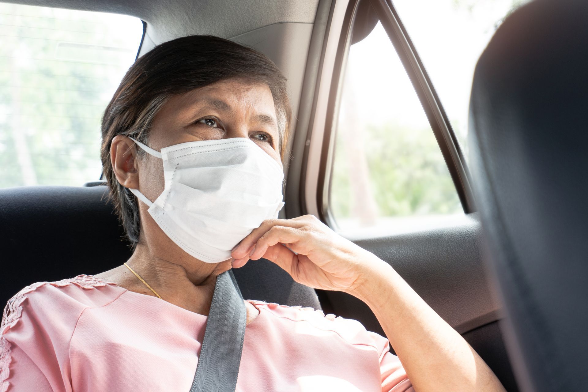 Une personne portant un masque et un t-shirt rose est assise dans une voiture, sa ceinture de sécurité attachée, près de la fenêtre.
