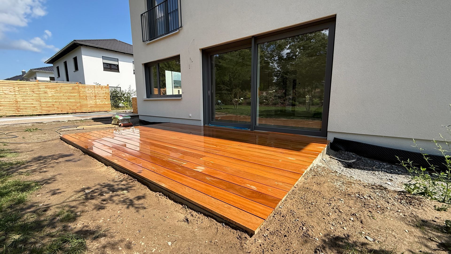 Eine Holzterrasse, die an ein helles Haus mit großen Fenstern angrenzt und in einem Garten liegt.