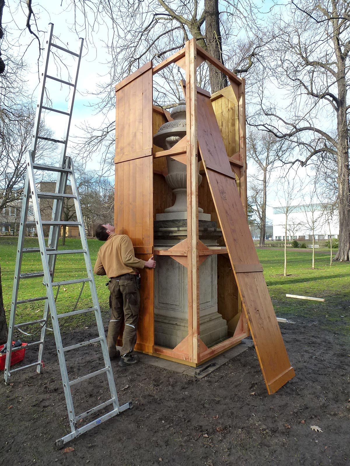 Ein Mann befestigt in einer Parkanlage Holzpaneele um ein Steinmonument; in der Nähe befindet sich eine Leiter.