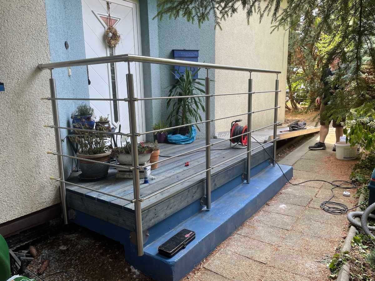 Edelstahlgeländer auf einer erhöhten Veranda mit grauem Bodenbelag, blauen Zierelementen und Pflanzen.