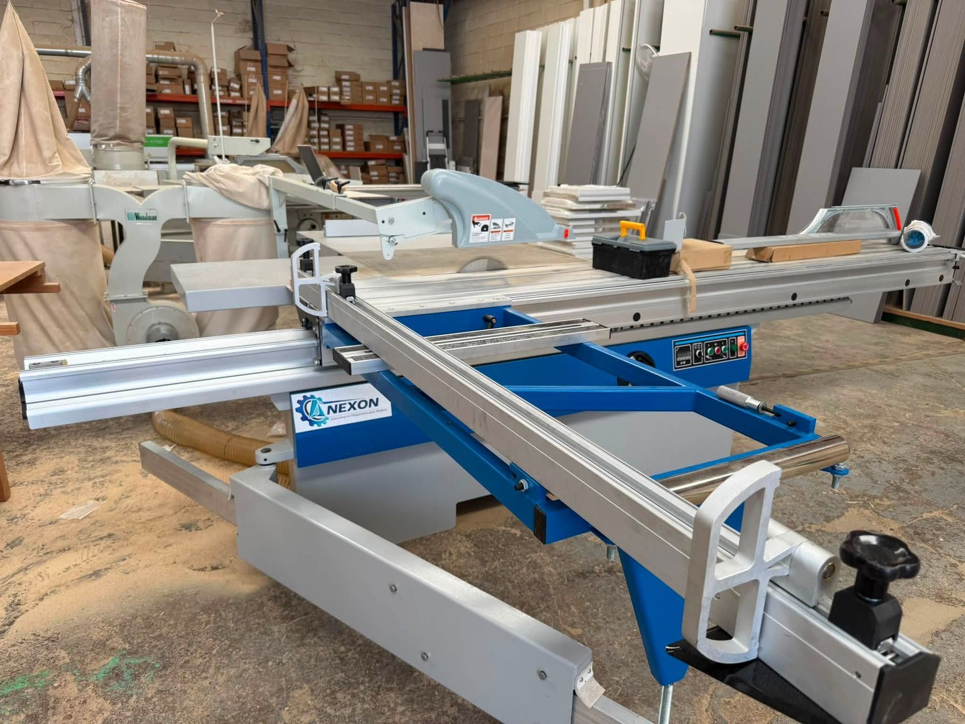 Sierra de mesa industrial con base azul y rieles plateados en un taller, rodeada de madera y herramientas