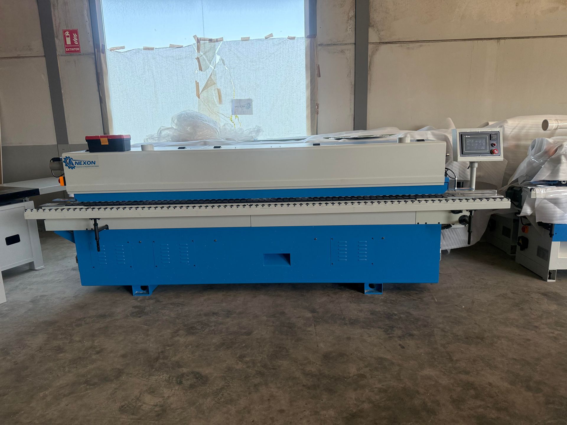 Máquina industrial azul en un taller, con la parte superior blanca y un panel de control cerca de una ventana.