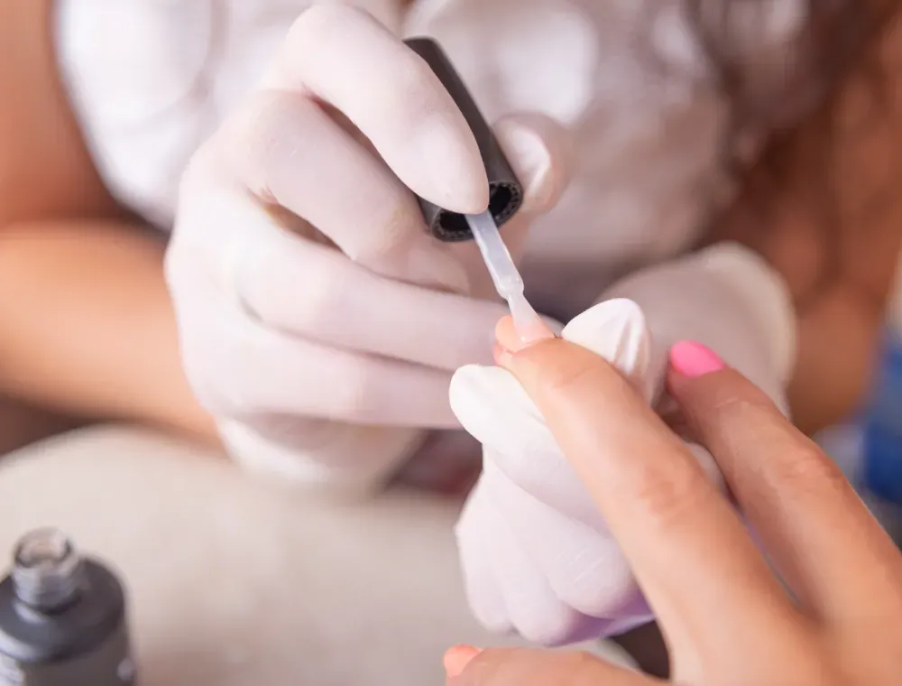 Manicurista cortando las uñas de una clienta con un cortaúñas. Se enfocan las manos, con guantes negros.
