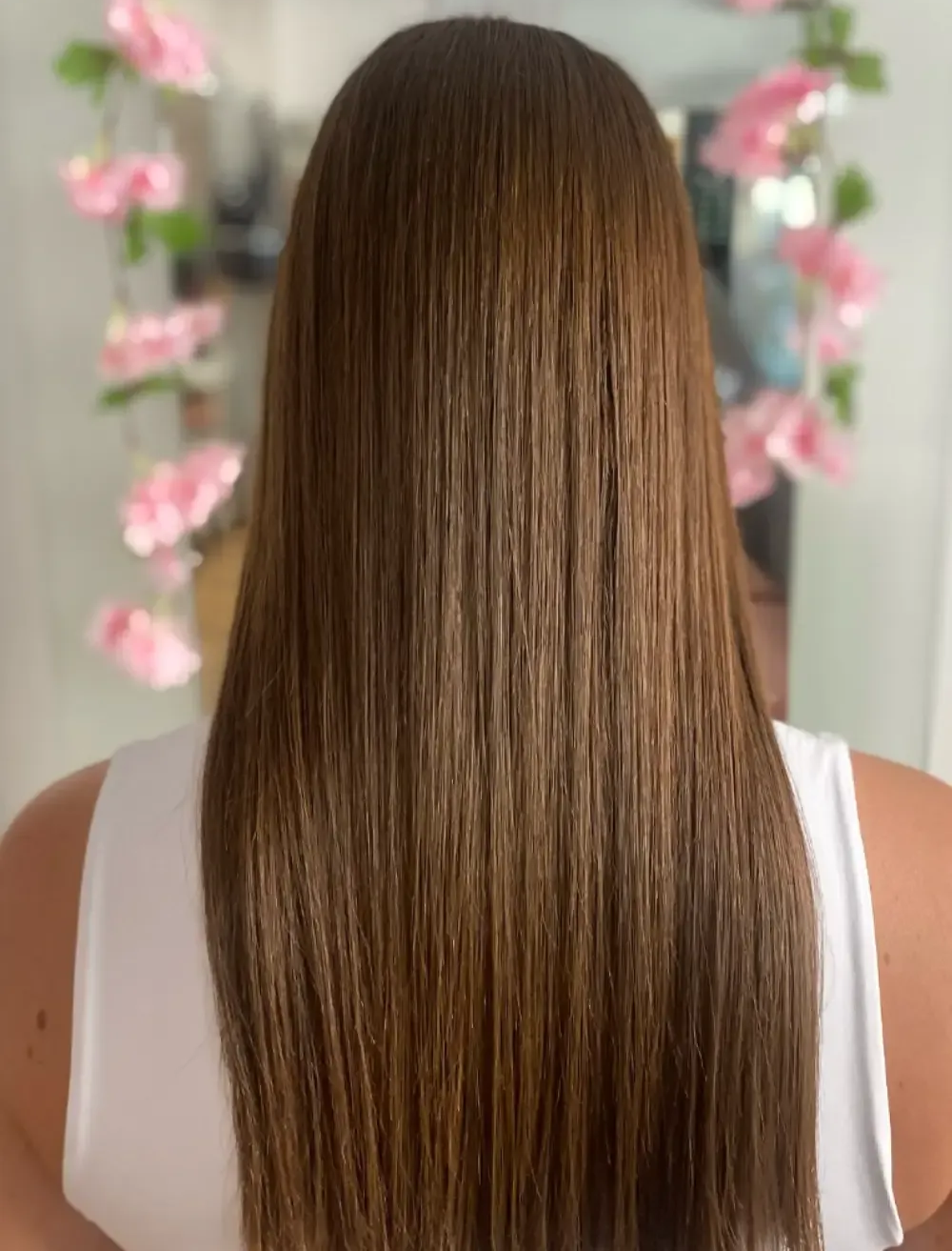 Mujer con cabello castaño largo y liso, vista desde atrás, en un salón, con flores rosas en el fondo.