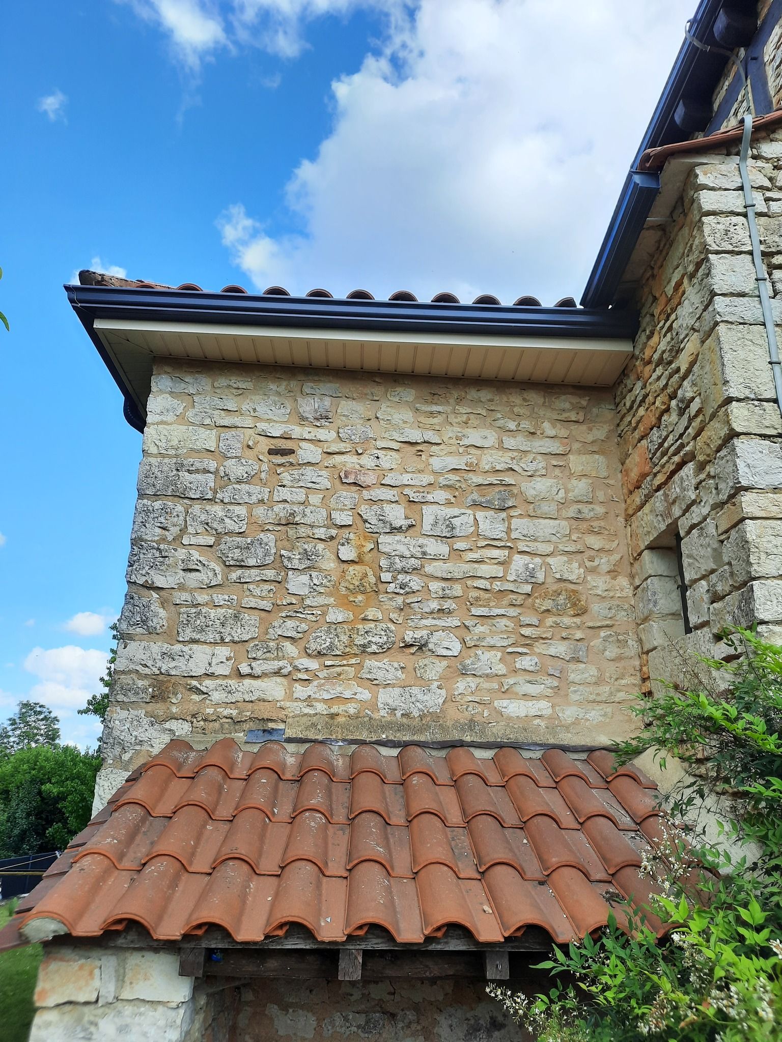 Mur d'un bâtiment en pierre avec un toit de tuiles rouges sous un ciel bleu éclatant et des nuages