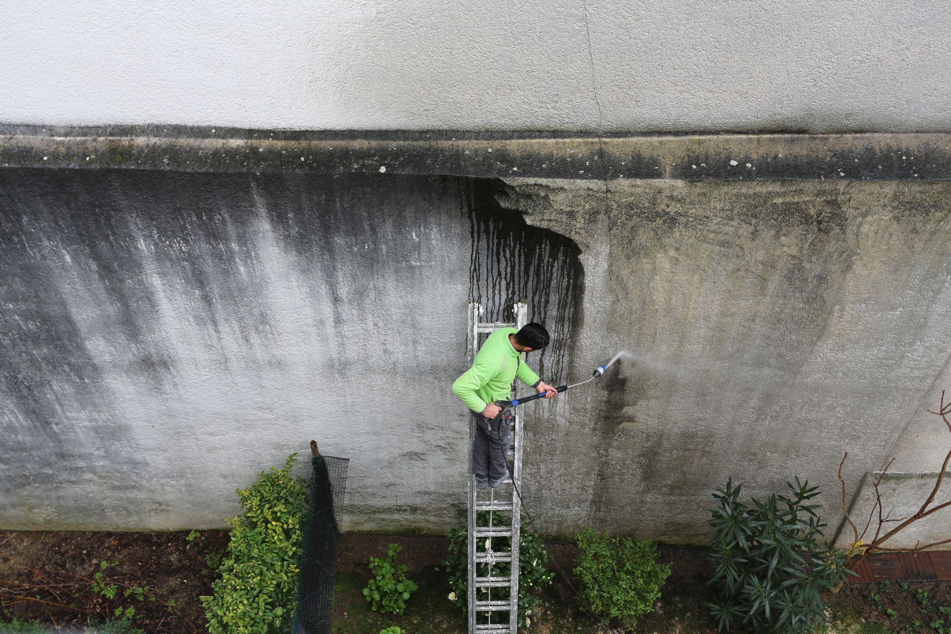 Un ouvrier portant une veste vert fluo utilise une échelle pour nettoyer un grand mur en béton.