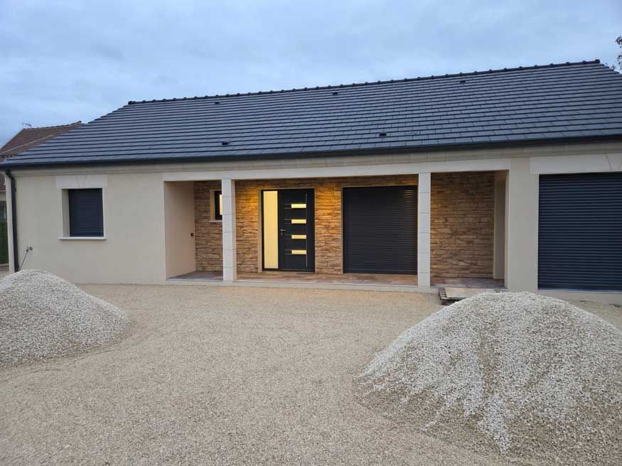 Vue extérieure d'une maison neuve avec des murs beige clair, un toit et des portes de garage noirs, et une allée en gravier.