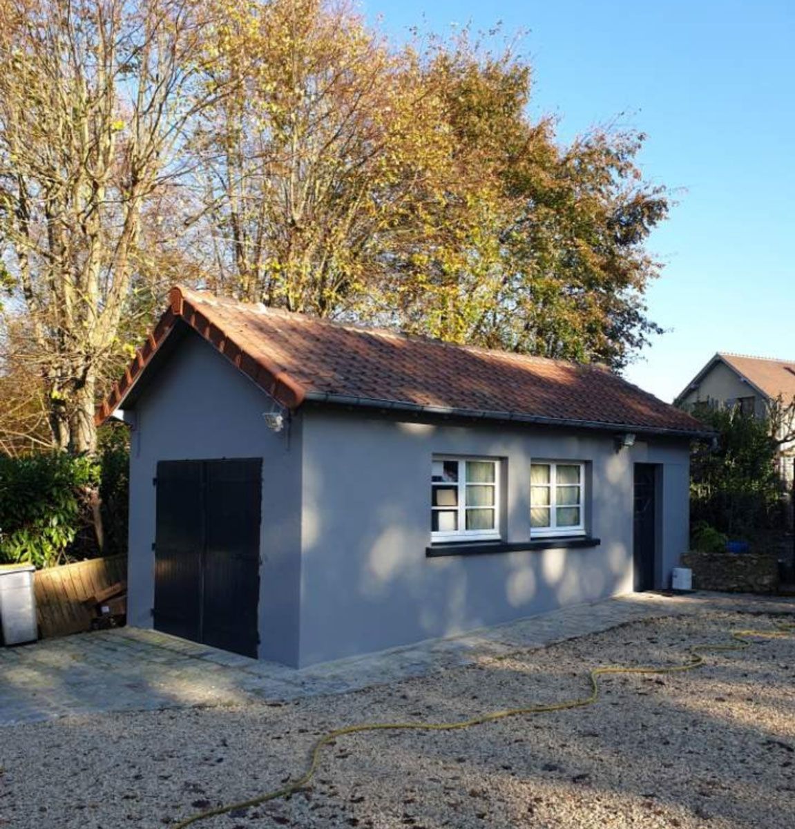 Garage peint en gris avec toit de tuiles rouges, deux fenêtres et portes de garage noires, dans une cour en gravier.