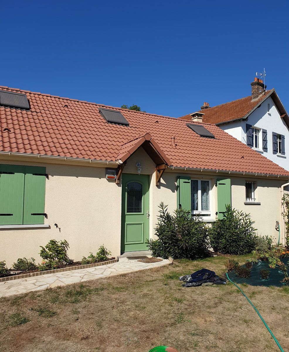 Un cottage au toit de tuiles rouges, à la porte et aux volets verts, par une journée ensoleillée. Un bâtiment blanc de deux étages se dresse à l'arrière-plan.