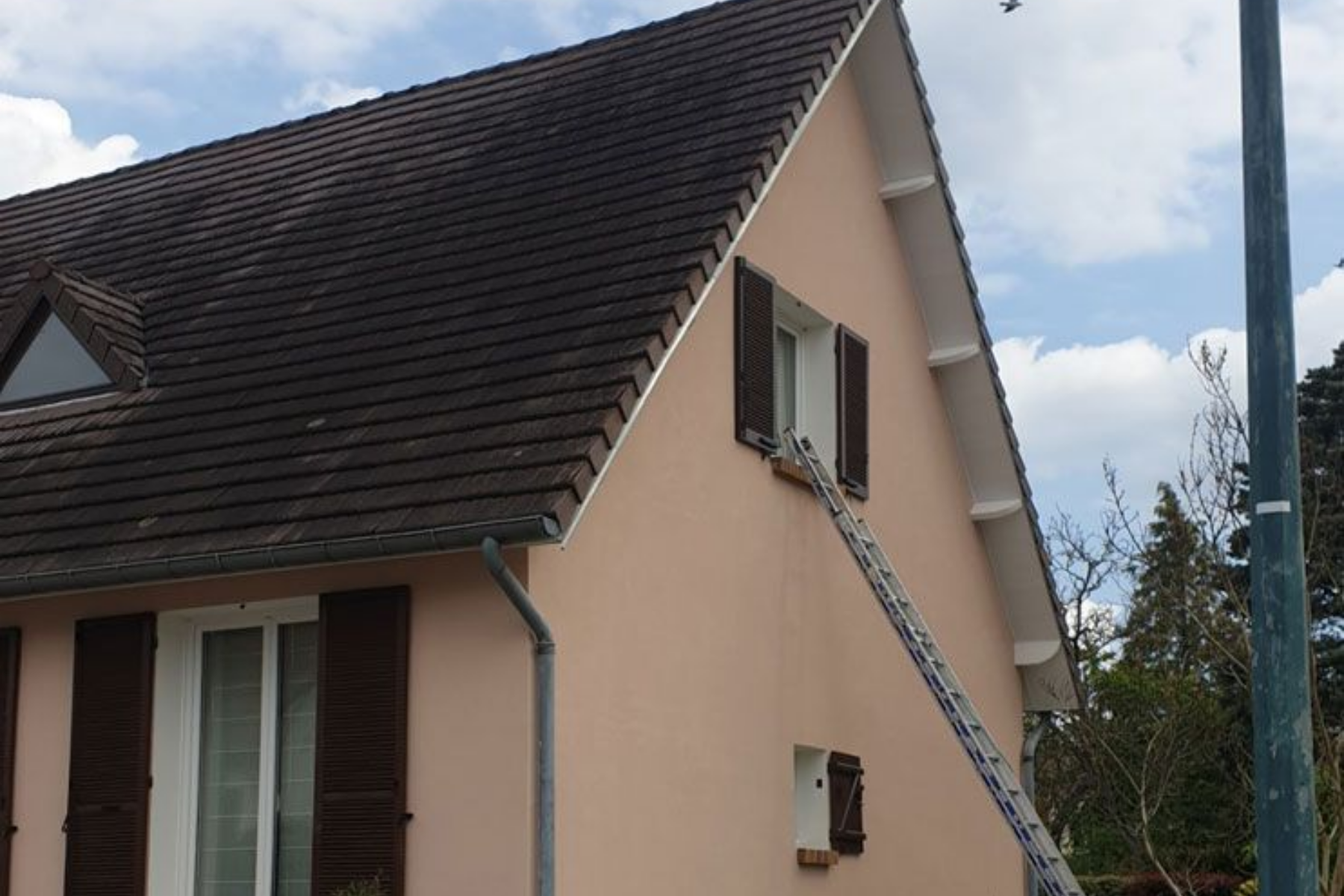 Maison rose avec toit et volets marron ; une échelle permet d'accéder à une fenêtre.