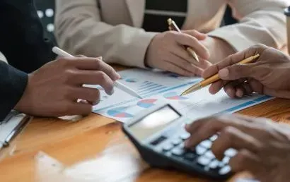 A group of people are sitting at a table using a calculator and a pen.