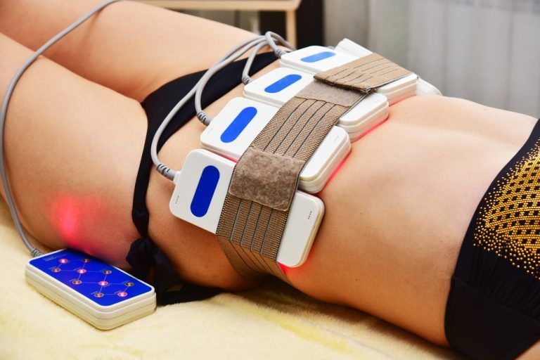Une personne allongée sur un lit avec un corset lombaire blanc et bleu et un téléphone allumé à proximité.