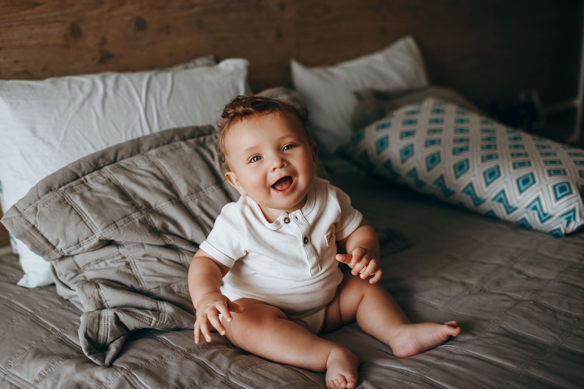 Bébé souriant assis sur un lit avec des draps gris et des oreillers à motifs.