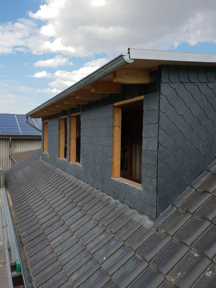 Dachgaube mit Schieferplatten und Holzrahmenfenstern, graue Dachschindeln und bewölkter Himmel im Hintergrund.
