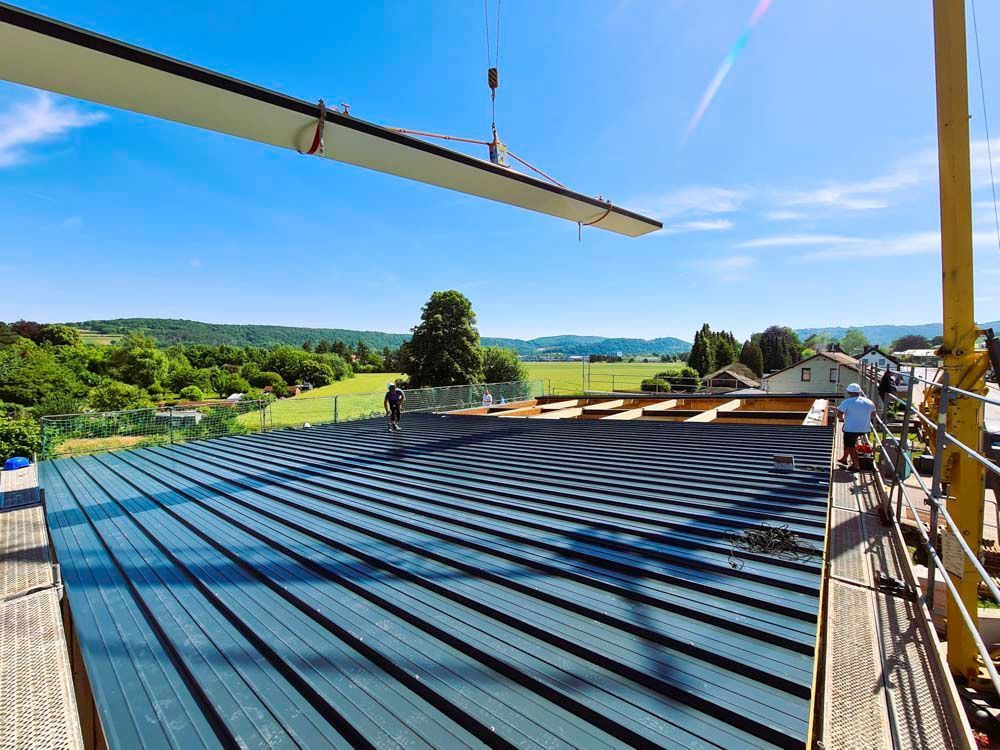Ein Kran hebt einen langen, hellen Träger über ein dunkelblaues Wellblechdach; Bauarbeiter auf dem Dach, im Hintergrund grüne Landschaft.