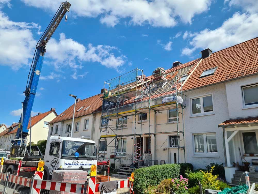 Bauarbeiter heben einen langen, rechteckigen Träger unter blauem Himmel auf ein blaues Wellblechdach.