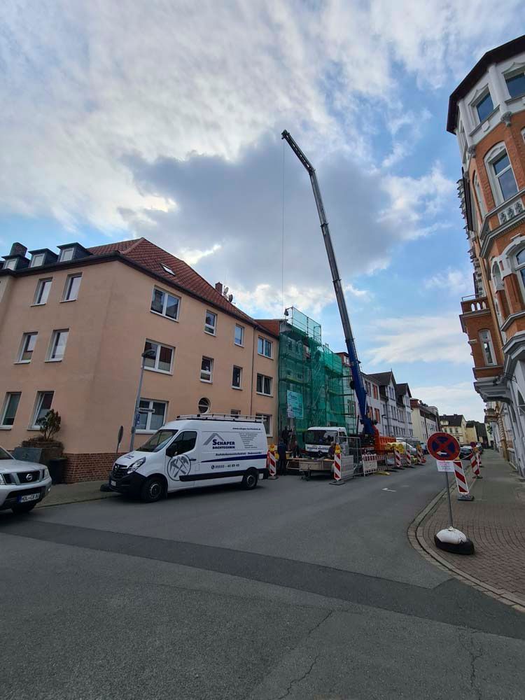 Ein Kran steht in der Stadtstraße und hebt Gegenstände zu einem mit grünen Netzen verhüllten Gebäude. Ein Lieferwagen steht geparkt, die Straße ist gesperrt, blauer Himmel.