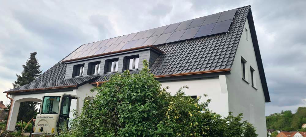 Weißes Haus mit Solarpaneelen auf dem Dach und bewölktem Himmel. Ein kleines Baufahrzeug steht in der Nähe des Hauses.