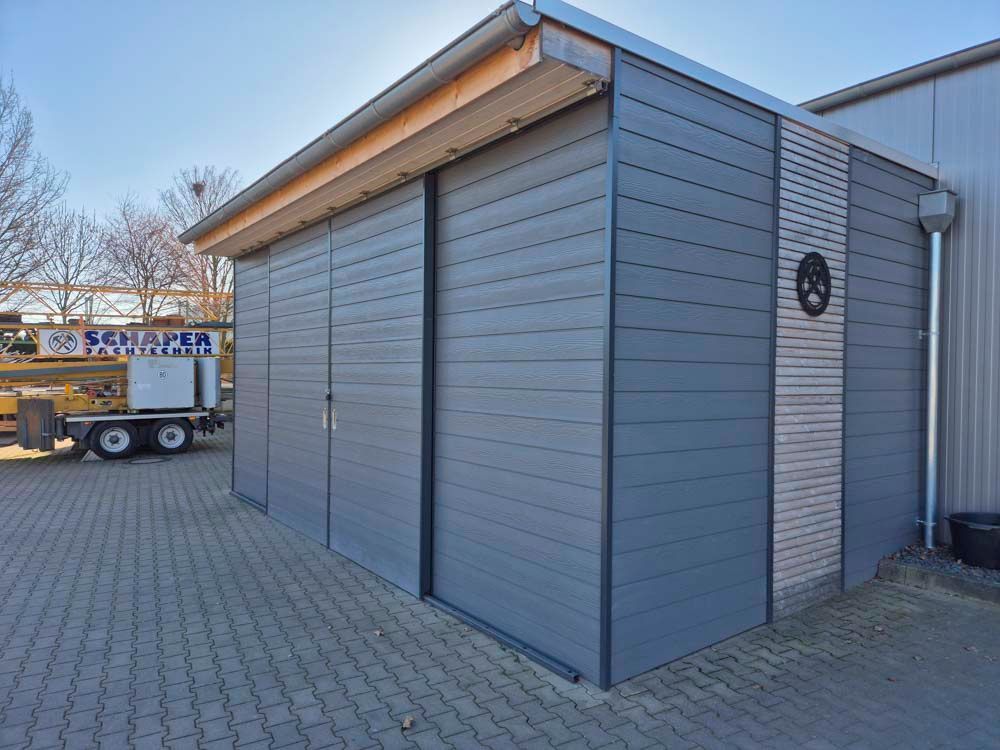 Grauer Geräteschuppen mit Schiebetüren, Backsteinpflaster und blauem Himmel.