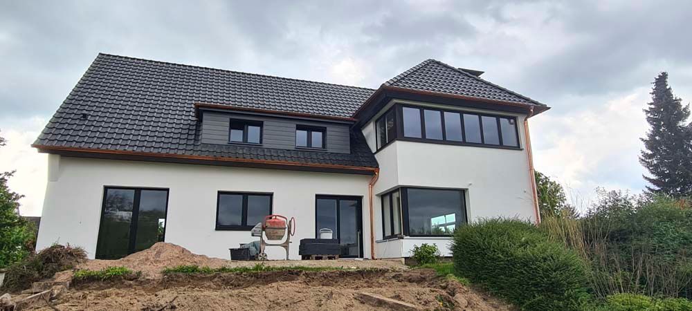 Weißes Haus mit dunklem Dach und Zierelementen unter bewölktem Himmel. Gartenarbeiten im Gange.