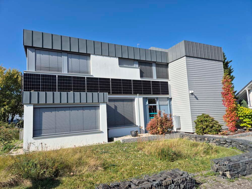 Zweistöckiges Gebäude mit Solarpaneelen und grauer Fassade. Strahlend blauer Himmel, Rasen und Büsche davor.
