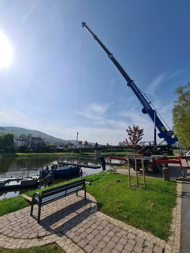 Ein Kran hebt ein Docksegment an einem Flussufer an, auf dem sich Boote, Menschen und eine Bank befinden.