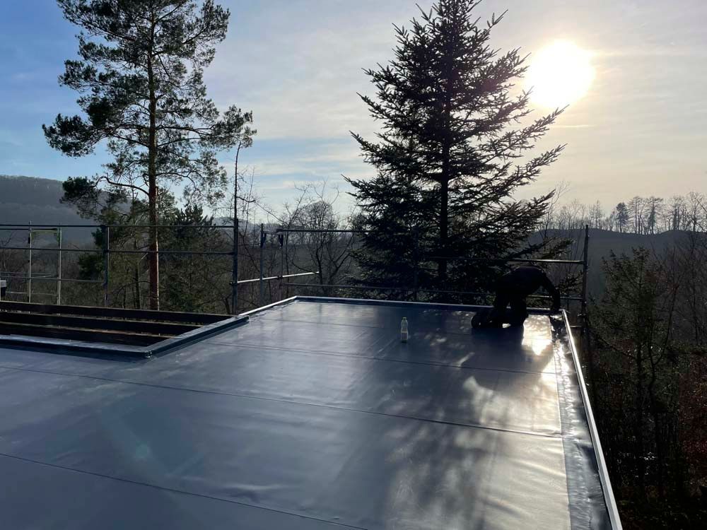 Eine Person arbeitet an einem sonnigen Tag auf einem Flachdach, umgeben von Bäumen.