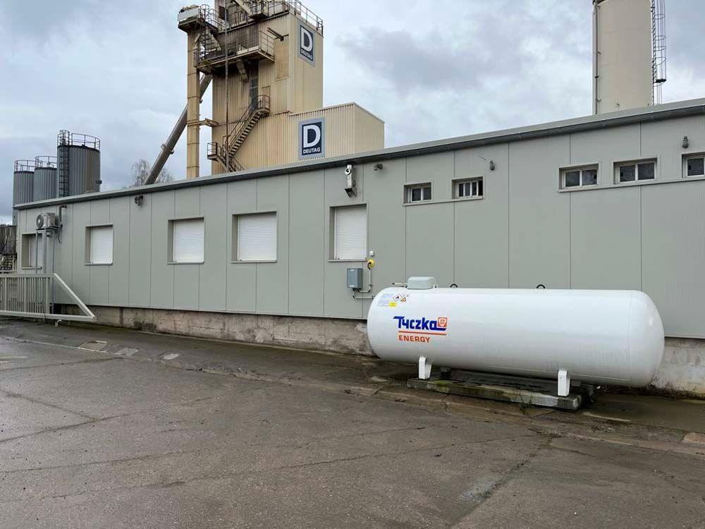 Ein weißer Propangastank steht vor einem hellgrauen Industriegebäude; im Hintergrund sind Silos zu sehen. Bewölkter Tag.