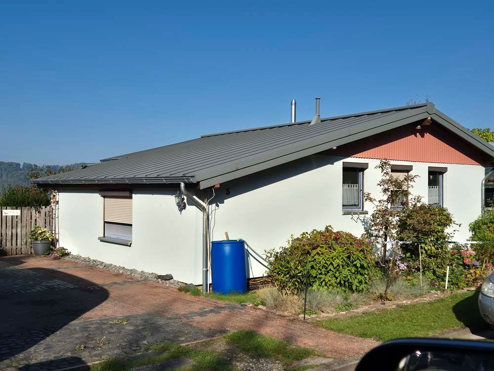 Weißes Haus mit grauem Dach, blauem Wasserfass und kleinem Vorgarten.